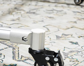 May include: Close-up of a silver metal furniture leg with a black caster wheel. The wheel has a white bearing and a black housing. The leg is attached to a white cylindrical piece. The background is a patterned rug.