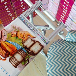 May include: A metal embroidery frame holding a piece of fabric with a cross-stitch pattern of a golden dog and brown luggage. The frame features adjustable silver bars and a color-coded legend strip along the side for thread selection.