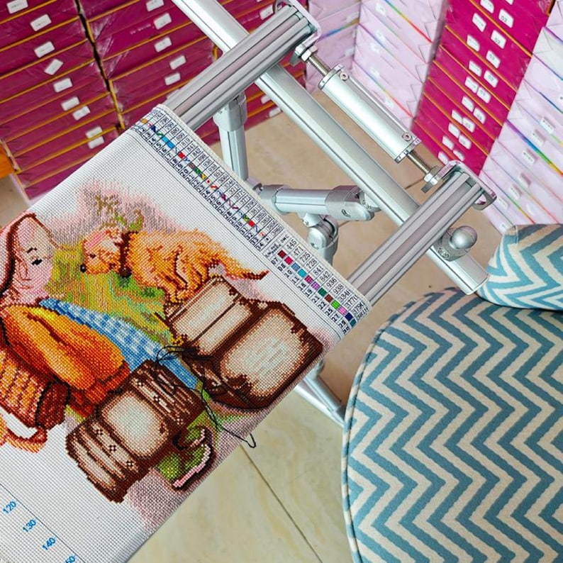 May include: A metal embroidery frame holding a piece of fabric with a cross-stitch pattern of a golden dog and brown luggage. The frame features adjustable silver bars and a color-coded legend strip along the side for thread selection.