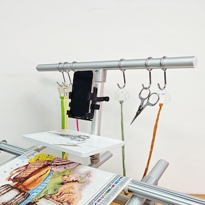 May include: A silver-colored craft stand with a white tray, a phone holder, and hooks for hanging tools. The stand holds a cross-stitch project with a colorful design. Scissors and thread are also visible.