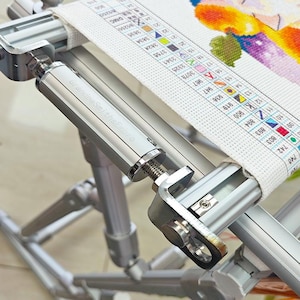 May include: A silver metal embroidery frame stand with adjustable tension knobs and a cylindrical component labeled GrandpaSmithsEmbro. The frame holds a piece of white fabric featuring a printed color-coded cross-stitch pattern chart.