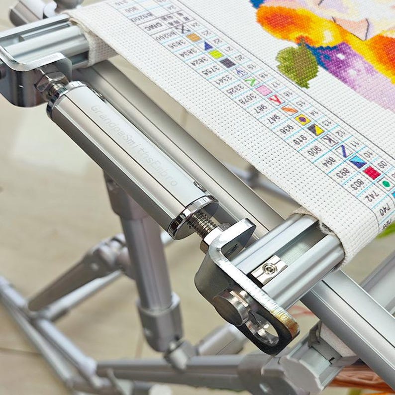 May include: A silver metal embroidery frame stand with adjustable tension knobs and a cylindrical component labeled GrandpaSmithsEmbro. The frame holds a piece of white fabric featuring a printed color-coded cross-stitch pattern chart.