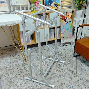 May include: A silver metal floor stand for embroidery or needlework projects, featuring an adjustable frame with multiple articulated joints, horizontal support bars, and a sturdy base with white end caps for stability during craft work.