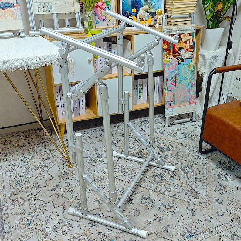 May include: A silver metal floor stand for embroidery or needlework projects, featuring an adjustable frame with multiple articulated joints, horizontal support bars, and a sturdy base with white end caps for stability during craft work.