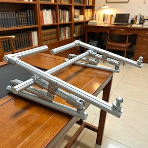 May include: A silver-colored adjustable laptop stand constructed from metal bars with multiple hinged joints. The frame features a rectangular base and an elevated support platform with two small rounded stoppers at the front edge to hold a device.