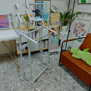 May include: A silver-colored, multi-jointed metal floor stand designed for embroidery frames. The structure features adjustable arms, a central horizontal support bar, and a small flat platform with a black clamp attachment for holding devices.