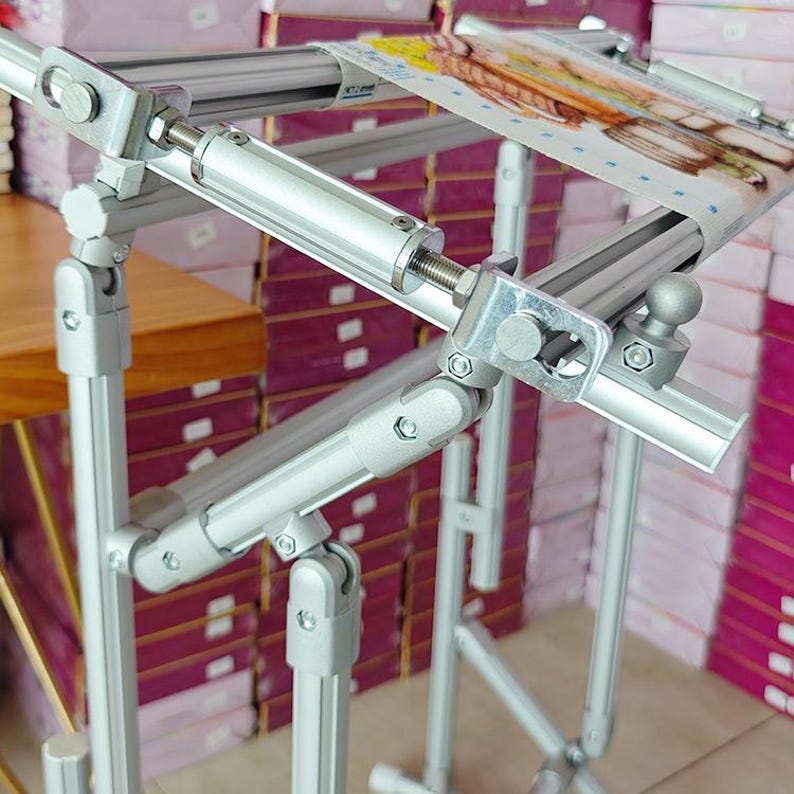 May include: A silver metal embroidery frame stand featuring adjustable aluminum bars and joints. The frame holds a piece of fabric with a printed design, secured by metal clips and tension screws, designed for hands-free needlework projects.