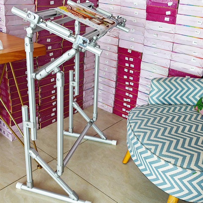 May include: A silver metal floor-standing embroidery frame with adjustable joints and a rectangular top bar. The frame holds a piece of fabric with a printed design, featuring multiple cylindrical support rods and a sturdy base structure.