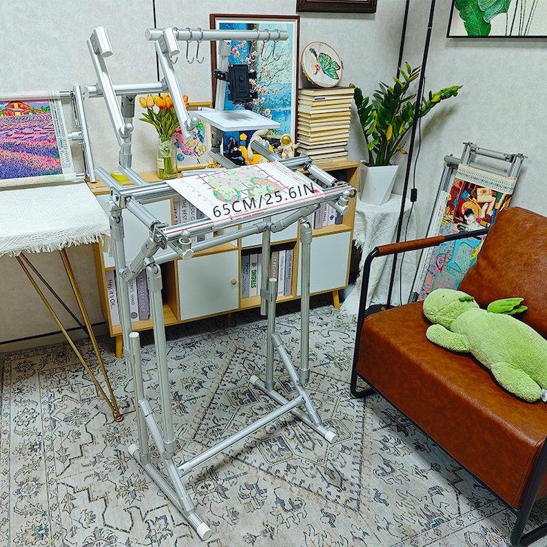 May include: A silver metal floor-standing embroidery frame with adjustable joints and a horizontal bar featuring the text 65CM/25.6IN. The frame includes a small white shelf, multiple metal hooks, and a black phone holder attachment for crafting.