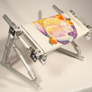 May include: A silver metal embroidery frame stand with adjustable joints and tension knobs, holding a piece of off-white fabric featuring a colorful cross-stitch design of a sleeping cat surrounded by orange fruit and purple circular patterns.