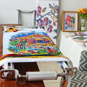 Puede incluir: Un bastidor de punto de cruz plateado con un diseño de punto de cruz colorido de una casa con un jardín. El bastidor tiene una perilla para ajustar la tensión de la tela.