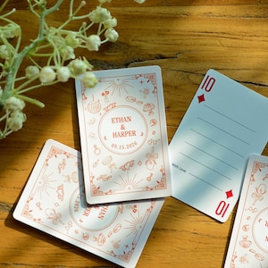 May include: White playing cards with red decorative borders and illustrations. One card features the number 10 and the suit of diamonds. Other cards display the names "Ethan & Harper" and the date "09.15.2026".