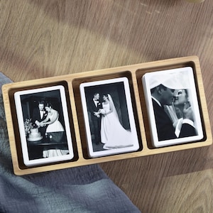 May include: A bamboo tray with three compartments, each holding a stack of black and white photographs. The photos depict wedding scenes, including a couple cutting a cake and a couple kissing. The tray sits on a wooden surface.