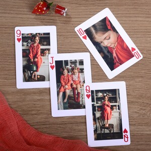 May include: Four playing cards with photos of children in red outfits. The cards are arranged on a wooden surface with a red fabric and decorative glass strawberry and gift box. The cards are the 9 of hearts, Jack of hearts, 10 of hearts, and Queen of hearts.
