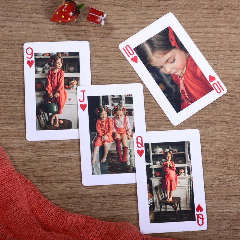 May include: Four playing cards with photos of children in red outfits. The cards are arranged on a wooden surface with a red fabric and decorative glass strawberry and gift box. The cards are the 9 of hearts, Jack of hearts, 10 of hearts, and Queen of hearts.