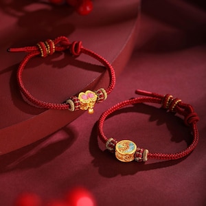 Puede incluir: Dos pulseras de cordón rojo con detalles dorados y abalorios decorativos. Una pulsera presenta un dije de flor con detalles rosas, la otra un dije circular con detalles azules y verdes. Las pulseras se muestran sobre una superficie roja.