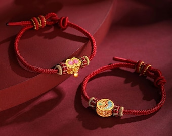 May include: Two red cord bracelets with gold accents and decorative charms. One bracelet features a flower charm with pink accents, while the other has a circular charm with blue and green details. The bracelets are displayed on a red surface.