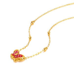 Puede incluir: Un collar dorado con un colgante de mariposa adornado con piedras preciosas rojas. La mariposa es el elemento central, suspendido de una fina cadena con pequeñas cuentas decorativas. El fondo es blanco.