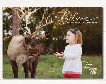 May include: A young girl in a white shirt and red pants smiles at a reindeer with antlers. The text "Believe in the magic of Christmas" is written in cursive above the girl and reindeer. The text "Love, Jordan, Megan & Kate" is written in cursive below the girl and reindeer.