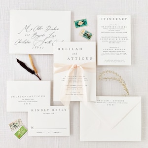 May include: A wedding stationery set featuring white paper cards and envelopes with black serif and script typography. The central invitation is tied with a pale pink ribbon. Included are an itinerary, reply card, and vintage postage stamps.