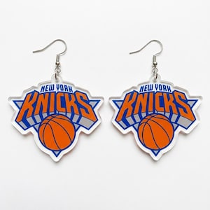 May include: A pair of dangle earrings featuring the New York Knicks logo. Each earring is a clear acrylic cutout shaped like the team emblem, displaying the text NEW YORK KNICKS in blue and orange with a basketball graphic and silver metal hooks.