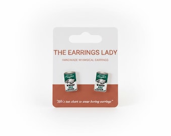 May include: A pair of stud earrings shaped like miniature soup cans with a green and white label featuring an eagle logo and the text Whoop Ass Soup. The earrings are displayed on a white and brown card labeled The Earrings Lady.