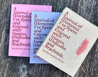 May include: Three small journals with the title "A Journal of Moments I've Spent and Enjoyed with Insects, Mollusks, and Arachnids." The journals are pink, blue, and tan, with red text and a small illustration.