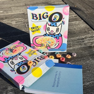May include: Two boxes of "BIG O" cereal with a cartoon character on the front, and the text "PRIZE INSIDE!". A small book with the text "Fuck Capitalism by Masturbating" is next to the cereal boxes. Colorful cereal pieces are scattered around.
