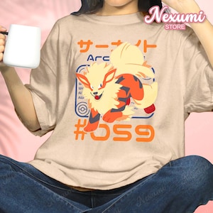 May include: Beige t-shirt featuring a vibrant cartoon illustration of a fiery creature with orange, yellow, and red accents. The design includes Japanese text, the word "Arc", and the number "059".