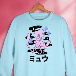 Könnte beinhalten: Hellblaues Sweatshirt mit einer rosa Cartoonfigur, blauen Augen, schwarzen Wolkenformen und blauen Sternakzenten. Japanischer Text ist unter der Figur aufgedruckt. Das Sweatshirt hängt an einem Holzkleiderbügel.