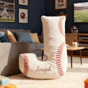Puede incluir: Un cojín blanco con forma de la letra "J", con temática de béisbol. Tiene costuras rojas y el nombre "Joseph" bordado. El cojín está en un salón, sobre una alfombra clara. La habitación tiene una temática deportiva.