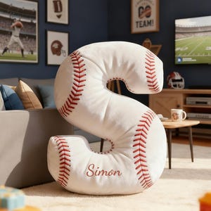 May include: A large, white, baseball-themed pillow in the shape of the letter "S". The pillow has red stitching and the name "Simon" embroidered in red. The pillow is on a white rug.