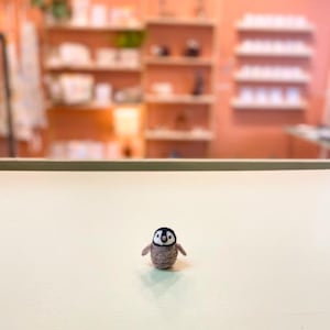 Puede incluir: Una pequeña figura de pingüino marrón y negro con el vientre blanco y los ojos negros. El pingüino está de pie sobre una superficie blanca.