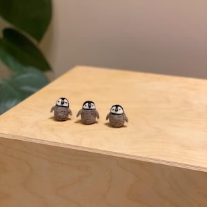 Puede incluir: Tres pingüinos de fieltro hechos a mano con cabezas negras y blancas y cuerpos grises, colocados sobre una superficie de madera. Los pingüinos son pequeños, con formas simples y redondeadas, y están alineados.