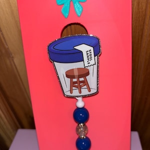 Può includere: Un portabadge di novità con un'immagine a cartoni animati di un contenitore per campioni di feci e la scritta "STOOL SAMPLE". Il portabadge ha un design a perline blu e bianche ed è attaccato a una carta rosa con un fiocco verde acqua.