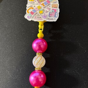 May include: A speech therapy themed badge reel with a white and colourful design. The badge reel has a yellow clip and a beaded strand with pink and white beads. The badge reel has the words "Speech Therapy" on it.