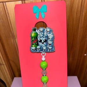 May include: A pink badge holder with a teal bow. The badge features a cartoon image of a Mountain Dew bottle and a cup of ice cubes with smiling faces. The lanyard has green and clear beads.