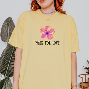 Puede incluir: Camiseta amarilla con un diseño floral rosa y morado y la frase "WALK FOR LOVE". La camiseta de manga corta tiene un corte relajado, ideal para un look casual.