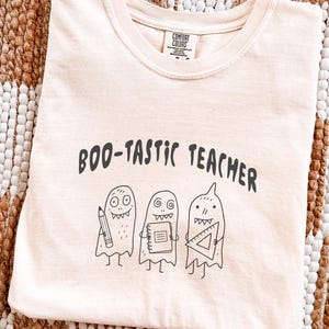 以下が含まれることがあります： 薄いピーチ色のTシャツで、黒い文字で「BOO-TASTIC TEACHER」と書かれ、その下に3体の幽霊の線画が描かれています。それぞれの幽霊は学用品を持っており、1体は鉛筆、1体はスパイラルノート、もう1体は三角定規を持っています。