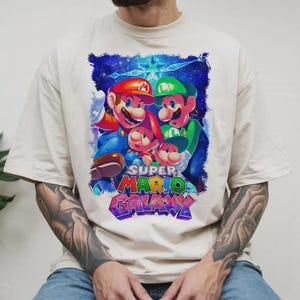 Puede incluir: Camiseta color crema con un gr&aacute;fico vibrante de Mario, Luigi y otros dos personajes del juego Super Mario Galaxy. El dise&ntilde;o incluye el logotipo del juego en texto colorido sobre un fondo estrellado.