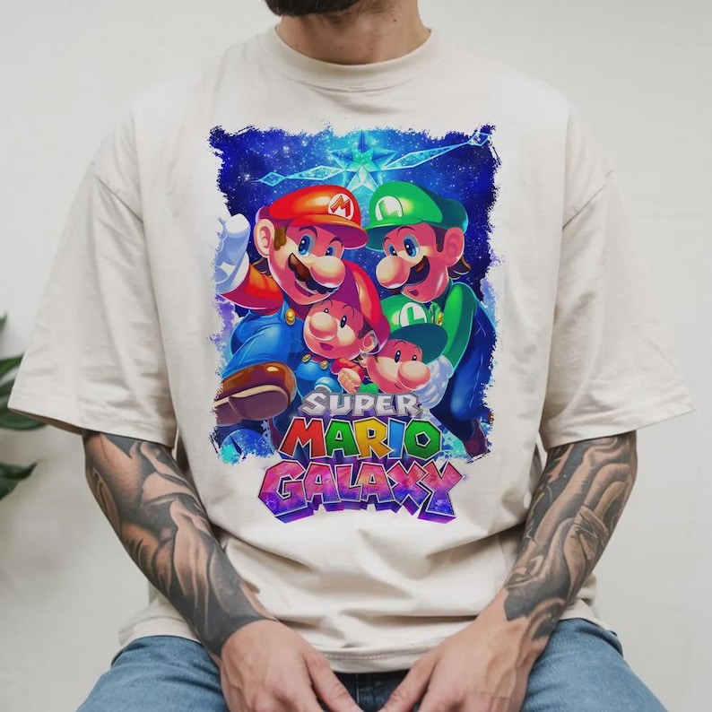 Puede incluir: Camiseta color crema con un gr&aacute;fico vibrante de Mario, Luigi y otros dos personajes del juego Super Mario Galaxy. El dise&ntilde;o incluye el logotipo del juego en texto colorido sobre un fondo estrellado.