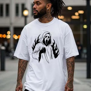 May include: White oversized t-shirt featuring a black and white graphic of a hooded figure holding a knife, surrounded by flames. The design is centered on the front of the shirt. The shirt has a crew neck and short sleeves.