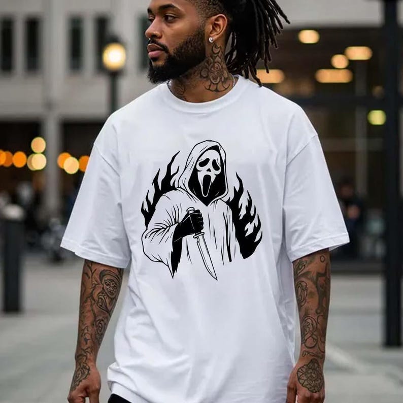 May include: White oversized t-shirt featuring a black and white graphic of a hooded figure holding a knife, surrounded by flames. The design is centered on the front of the shirt. The shirt has a crew neck and short sleeves.