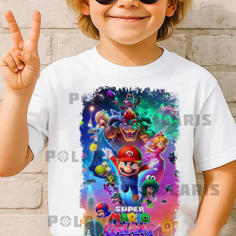 K&ouml;nnte beinhalten: Wei&szlig;es T-Shirt mit einer farbenfrohen Grafik von Charakteren aus Super Mario Galaxy. Das Design zeigt Mario, Prinzessin Peach, Bowser und andere Figuren. Der Text "SUPER MARIO GALAXY" ist unten zu sehen.
