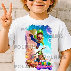 K&ouml;nnte beinhalten: Wei&szlig;es T-Shirt mit einer farbenfrohen Grafik von Luigi aus Super Mario Galaxy auf einem gr&uuml;nen Motorrad. Das Design umfasst einen blauen Himmel, Planeten und das Logo des Spiels in leuchtenden Farben. Das Kind tr&auml;gt eine Sonnenbrille und zeigt das Friedenszeichen.