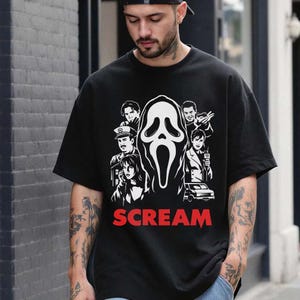 May include: Black t-shirt featuring a white and black graphic of the Scream mask and characters from the movie. The word "SCREAM" is printed in red below the graphic. A classic horror movie tee.