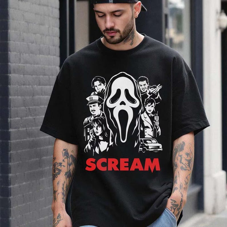 May include: Black t-shirt featuring a white and black graphic of the Scream mask and characters from the movie. The word "SCREAM" is printed in red below the graphic. A classic horror movie tee.