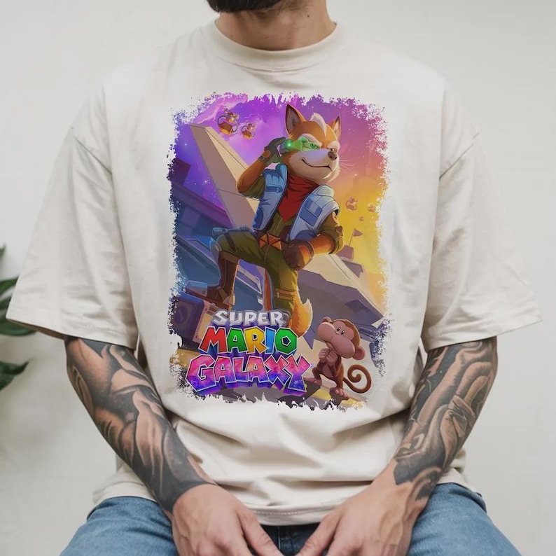 Puede incluir: Camiseta color crema con un gr&aacute;fico vibrante de un personaje parecido a un zorro con visera verde y un mono, del juego Super Mario Galaxy. El dise&ntilde;o incluye el logotipo del juego en texto colorido, sobre un fondo espacial.