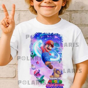 K&ouml;nnte beinhalten: Wei&szlig;es T-Shirt mit einem farbenfrohen Super Mario Galaxy-Motiv. Das Design zeigt Mario in seiner roten Kappe und blauen Latzhose, umgeben von Spielfiguren und dem Logo des Spiels. Das T-Shirt wird von einem Kind getragen.