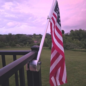 May include: A silver metal flag pole mounting bracket attached to a black railing. The bracket features a cylindrical base with a tightening knob and a diagonal sleeve holding a silver pole. The text reads: Flag, flag pole and railing not included.